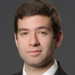A man with short dark hair wearing a white shirt and a dark suit jacket, posing in front of a plain dark background. He has a neutral facial expression and is looking directly at the camera.