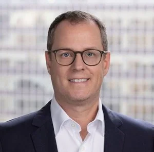 A man with short brown hair and glasses, wearing a white dress shirt and dark suit jacket, smiles at the camera. The background is blurred with a grid-like pattern.