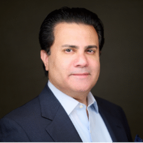 A man with dark hair, wearing a dark suit jacket and light blue shirt, is looking at the camera with a neutral expression against a dark background.