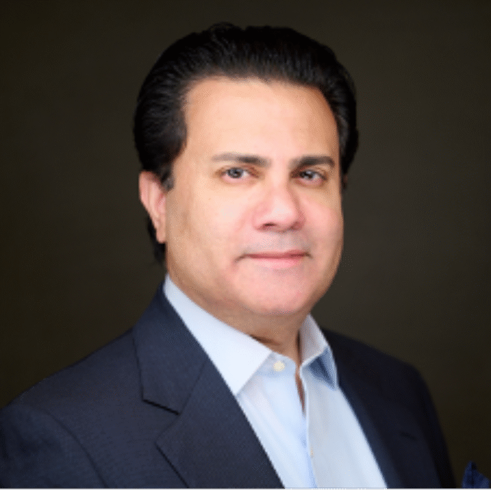 A man with dark hair, wearing a dark suit jacket and light blue shirt, is looking at the camera with a neutral expression against a dark background.