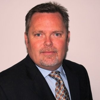 A middle-aged man with short brown hair and a goatee, wearing a dark suit, light blue shirt, and patterned tie, posing in front of a plain light background.