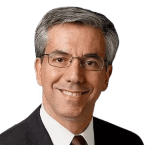 A man with short gray hair and glasses is smiling. He is wearing a dark suit, white shirt, and brown tie against a plain light background.