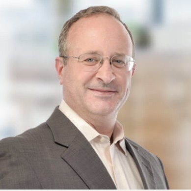 A middle-aged man with short light brown hair and glasses, wearing a gray suit jacket and light-colored shirt, smiling in front of a softly blurred indoor background.
