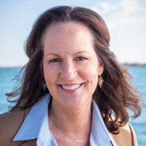 A woman with wavy brown hair smiles at the camera. She is wearing a light blue shirt, tan blazer, and gold starburst earrings, standing outdoors with blue water and a bright sky in the background.
