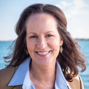 A woman with wavy brown hair smiles at the camera. She is wearing a light blue shirt, tan blazer, and gold starburst earrings, standing outdoors with blue water and a bright sky in the background.