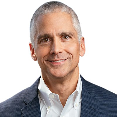 A middle-aged man with short gray hair, wearing a dark blue blazer and a white collared shirt, smiles warmly at the camera against a plain white background.