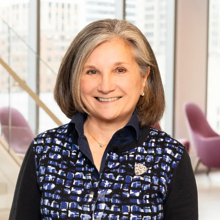A smiling woman with gray hair wearing a patterned vest over a dark shirt stands indoors in a modern office with large windows, a staircase, and two pink chairs in the background.