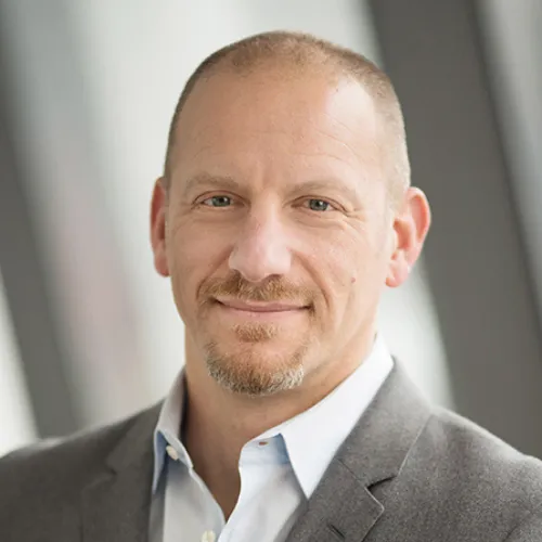 A man with light skin, short light brown hair, and a goatee, wearing a gray blazer and light blue shirt, smiles softly while standing in front of blurred windows.