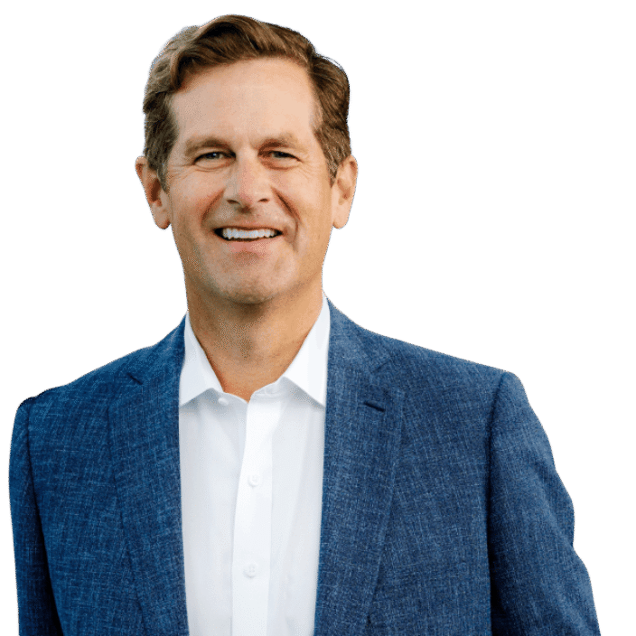 A man with short brown hair wearing a blue textured suit jacket over a white dress shirt stands smiling against a plain white background.