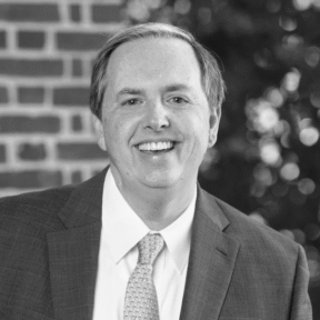 A man in a suit and tie smiles at the camera. The background features a brick wall on one side and blurred greenery on the other. The photo is in black and white.