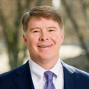A man with short light brown hair, wearing a dark blue suit, white shirt, and pink patterned tie, is smiling at the camera. The background is outdoors and blurred with trees.