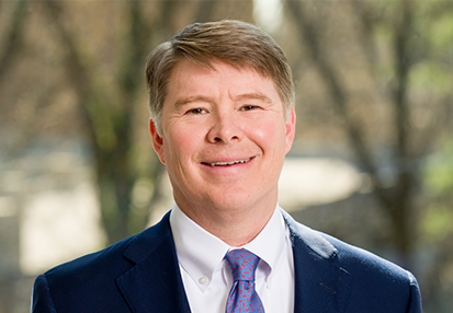 A man with short light brown hair, wearing a dark blue suit, white shirt, and pink patterned tie, is smiling at the camera. The background is outdoors and blurred with trees.