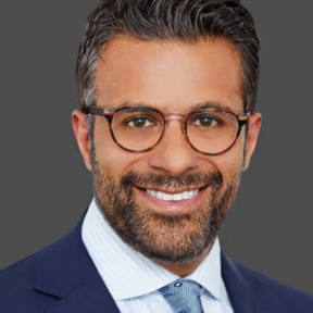 A man with short, dark hair, a beard, and glasses smiles while wearing a navy blue suit, light blue shirt, and patterned tie against a plain grey background.