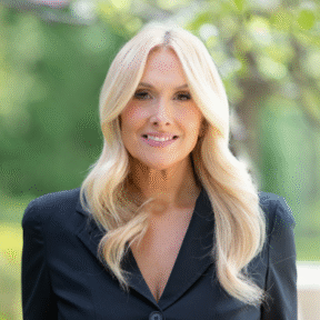 A woman with long, wavy blonde hair wearing a black blazer smiles while standing outdoors with greenery and soft sunlight in the background.