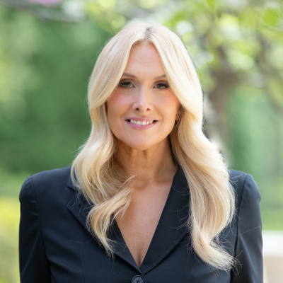 A woman with long, wavy blonde hair wearing a black blazer smiles while standing outdoors with greenery and soft sunlight in the background.