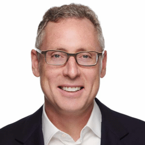 A man with short curly gray hair, wearing glasses, a white collared shirt, and a dark blazer smiles at the camera against a plain white background.