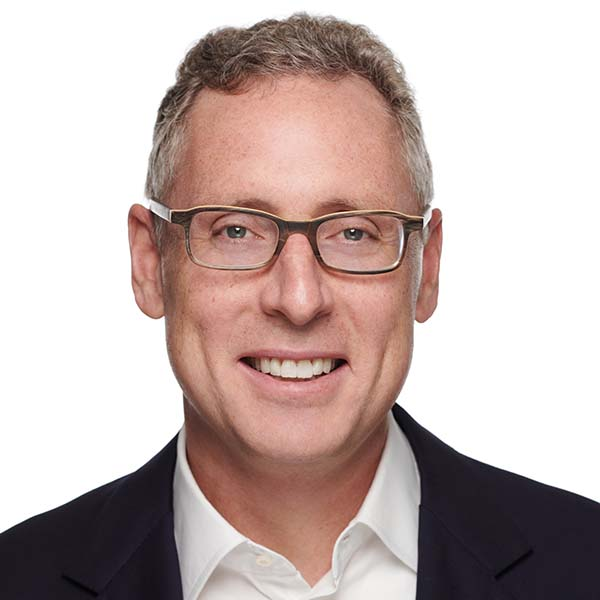 A man with short curly gray hair, wearing glasses, a white collared shirt, and a dark blazer smiles at the camera against a plain white background.