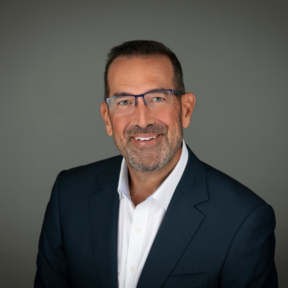 A middle-aged man with short dark hair, a beard, and glasses is smiling. He is wearing a dark suit jacket and white dress shirt, and is posed against a plain gray background.