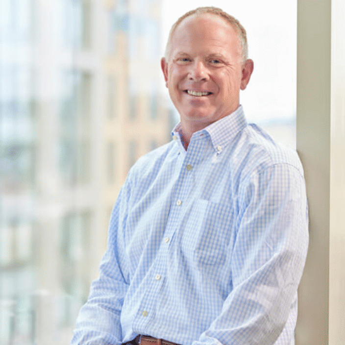 A middle-aged man in a light blue checkered shirt and dark pants sits on a window ledge, smiling at the camera. The background shows a bright, modern building interior with large windows.
