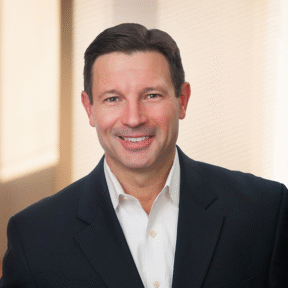 A man with short dark hair, wearing a dark suit jacket and white dress shirt, smiles at the camera. The background is softly lit with blurred vertical blinds and warm light.