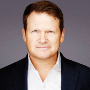 A man with light skin, short brown hair, and blue eyes is wearing a white shirt and dark blazer, smiling slightly while looking at the camera against a plain grey background.