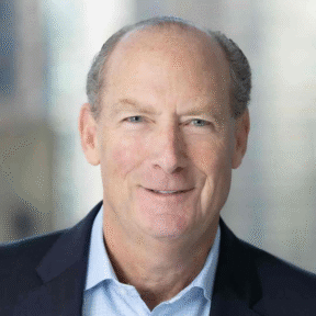 A middle-aged man with light skin, thinning gray hair, and blue eyes smiles at the camera. He is wearing a light blue dress shirt and a dark navy blazer, set against a softly blurred background.