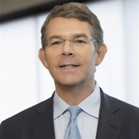 A middle-aged man with short brown hair, wearing glasses, a dark suit, a light blue shirt, and a light blue patterned tie, standing indoors in front of large windows with soft, diffused light.
