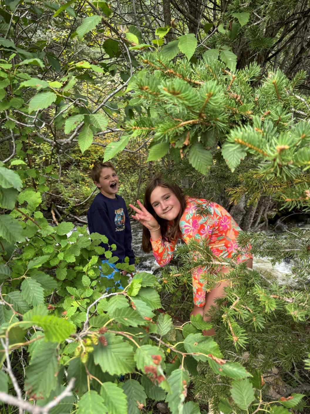 Two children, one in a dark shirt and another in a colorful floral top, smile and pose playfully among lush green trees and foliage near a creek in a forested area.
