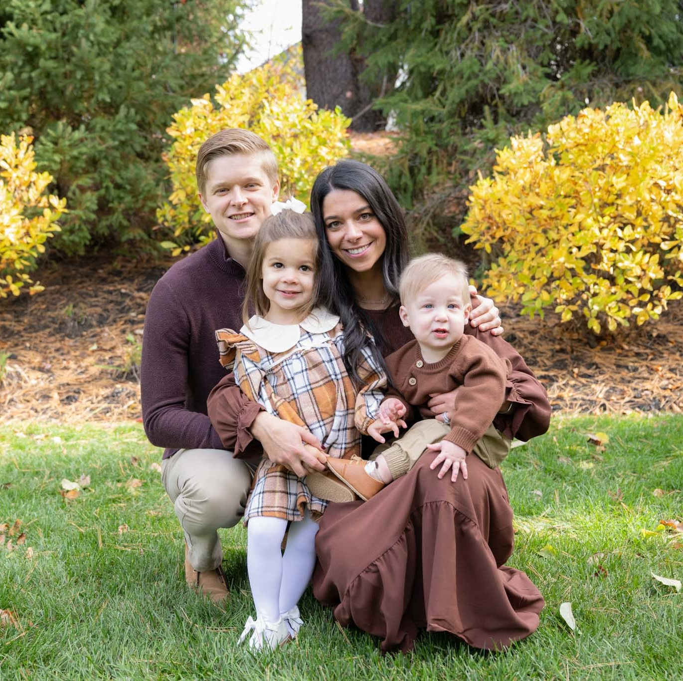 A family of four poses outdoors on green grass with autumn trees behind them. The parents kneel, smiling, with their two young children in front, all wearing coordinating brown and plaid outfits.