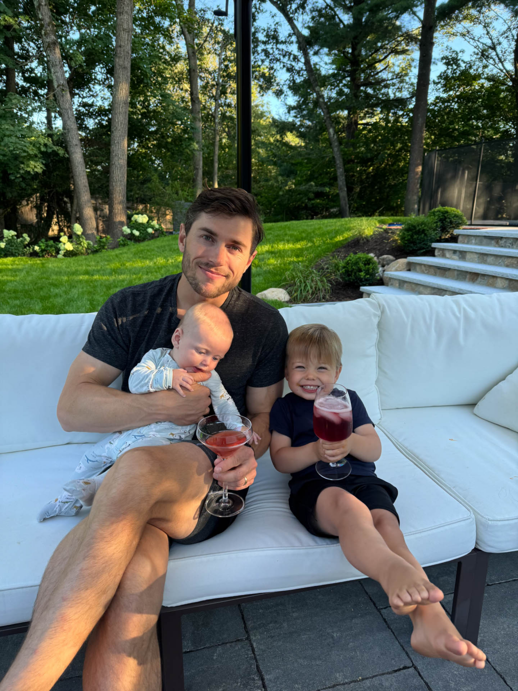 A man sits on an outdoor white couch holding a baby and a pink drink, with a young boy beside him smiling and holding a similar drink. Trees and greenery fill the background.