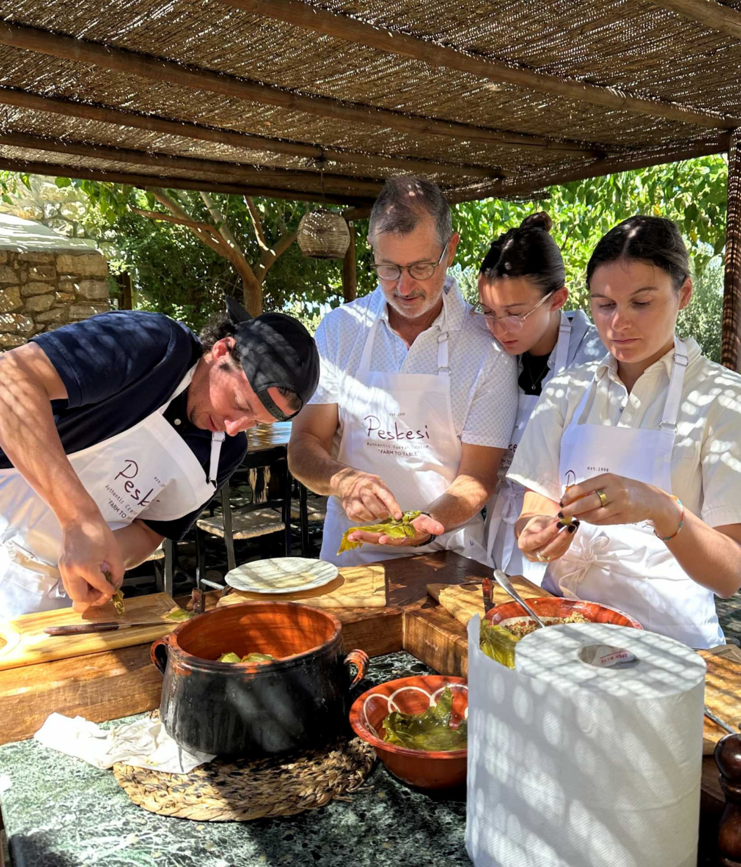 Four people wearing white aprons prepare food together at an outdoor table under a shaded canopy, surrounded by bowls of ingredients and greenery.
