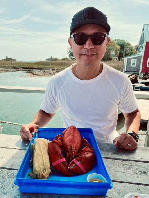 A man in sunglasses and a cap smiles while sitting outdoors by the water with a tray holding a cooked lobster, an ear of corn, and a cup of sauce.
