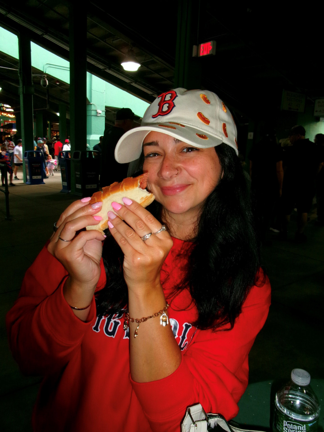 A woman wearing a Boston Red Sox cap and red sweatshirt smiles while holding a hot dog at an indoor stadium or concession area.