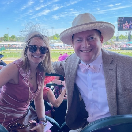 A smiling woman and man dressed in stylish, festive outfits and hats pose in stadium seats at a horse racetrack on a sunny day, with a crowd and racetrack visible in the background.