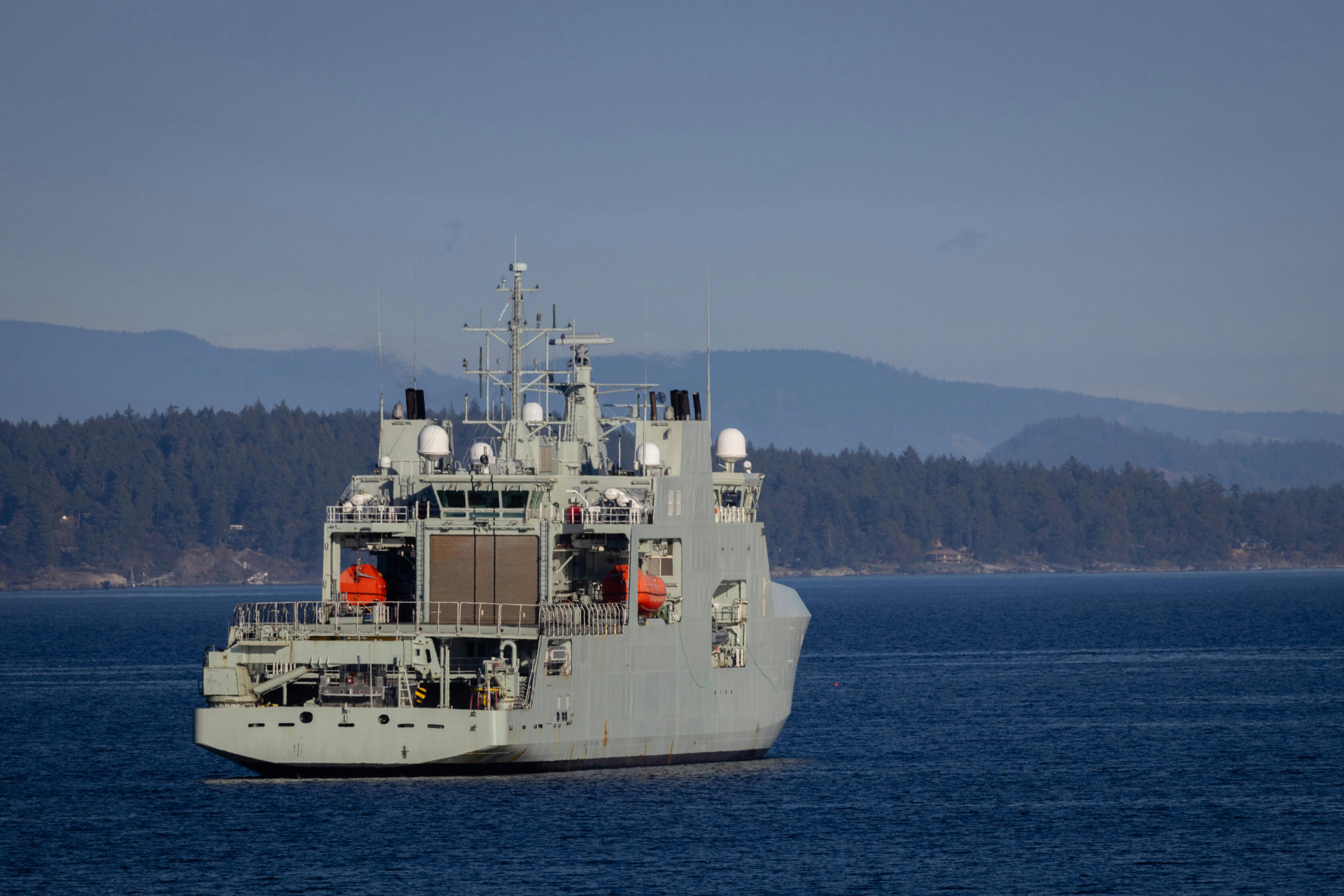 A large gray naval ship sails on calm blue water near a forested shoreline, with mountains in the background under a clear sky.