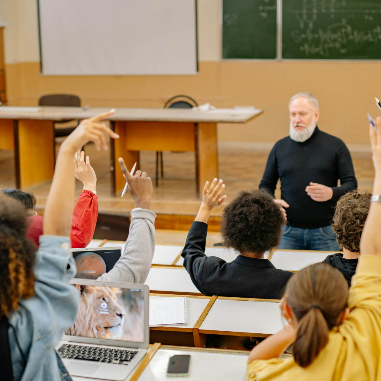 A professor stands at the front of a classroom, addressing students seated at desks. Several students have raised their hands, laptops are open, and behind the professor are chalkboards and a projector screen displaying a Carnegie Case Study.