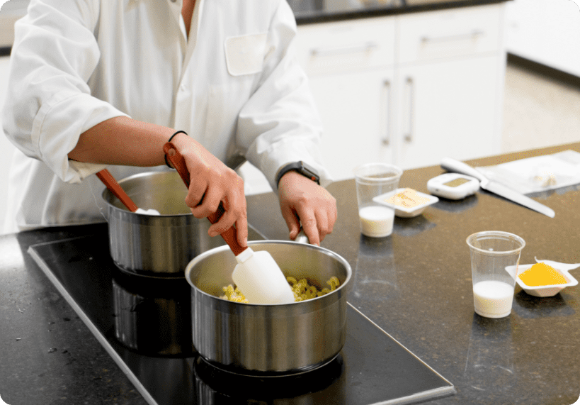 A person in a white lab coat stirs ingredients in a stainless steel pot on a stovetop, with containers of milk and spices arranged nearby—showcasing a behind-the-scenes glimpse into product development at Revela Foods.