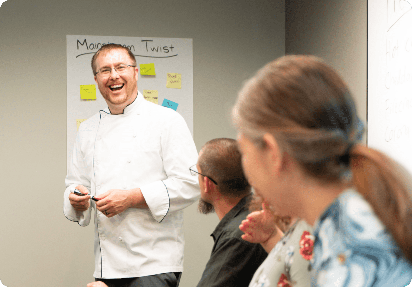 A smiling man in a white chef’s coat stands near a whiteboard, engaging with seated people in a Revela Foods case study meeting. Colorful sticky notes are on the wall behind him, and participants appear attentive and cheerful.