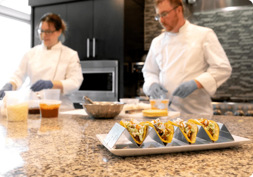 Two chefs in white uniforms and blue gloves prepare food in a modern kitchen for a Revela Foods case study, with a tray of four assembled tacos on the countertop and bowls of ingredients visible near the chefs.