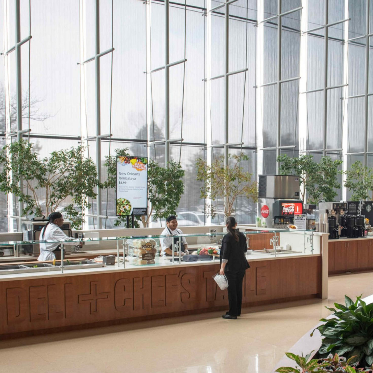 A modern cafeteria with large glass windows, food counters staffed by workers, and digital menu boards. As seen in the Continental Services Case Study, green plants add freshness to the bright, open space where people order or carry food.