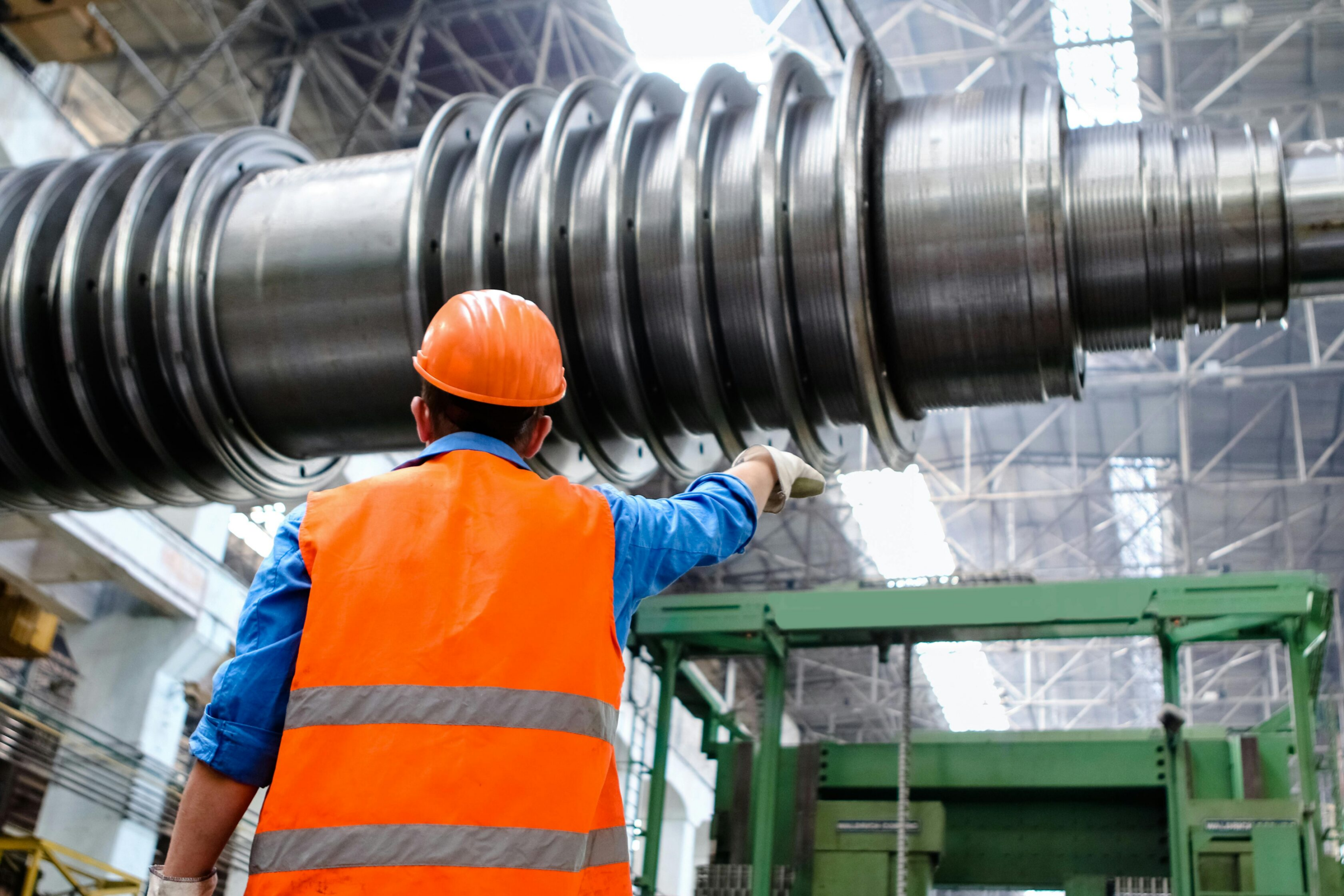 A worker in an orange safety vest and hard hat stands in a large industrial facility, pointing toward a massive, cylindrical metal machine part suspended from above—showcasing Our Difference in safety and precision.