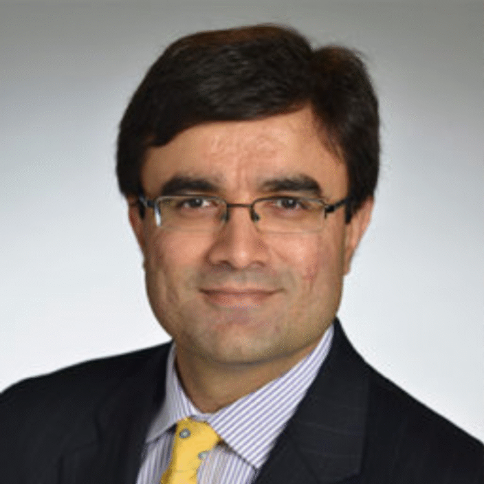 A man with short dark hair and glasses, dressed in a dark pinstripe suit, light striped shirt, and a yellow tie with blue dots, is posed in front of a plain light background—reflecting the professionalism of Sutton Place Strategies.
