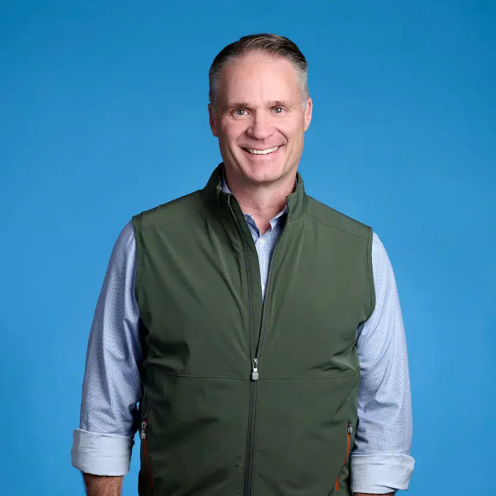 A man with short hair smiles at the camera, wearing a light blue shirt with rolled-up sleeves and a green zip-up vest. The bright blue background reflects his professional role at Incline Equity Partners in private equity investment.