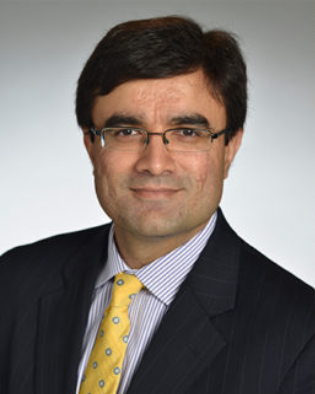 A man with short dark hair and glasses, dressed in a dark pinstripe suit, light striped shirt, and a yellow tie with blue dots, is posed in front of a plain light background—reflecting the professionalism of Sutton Place Strategies.