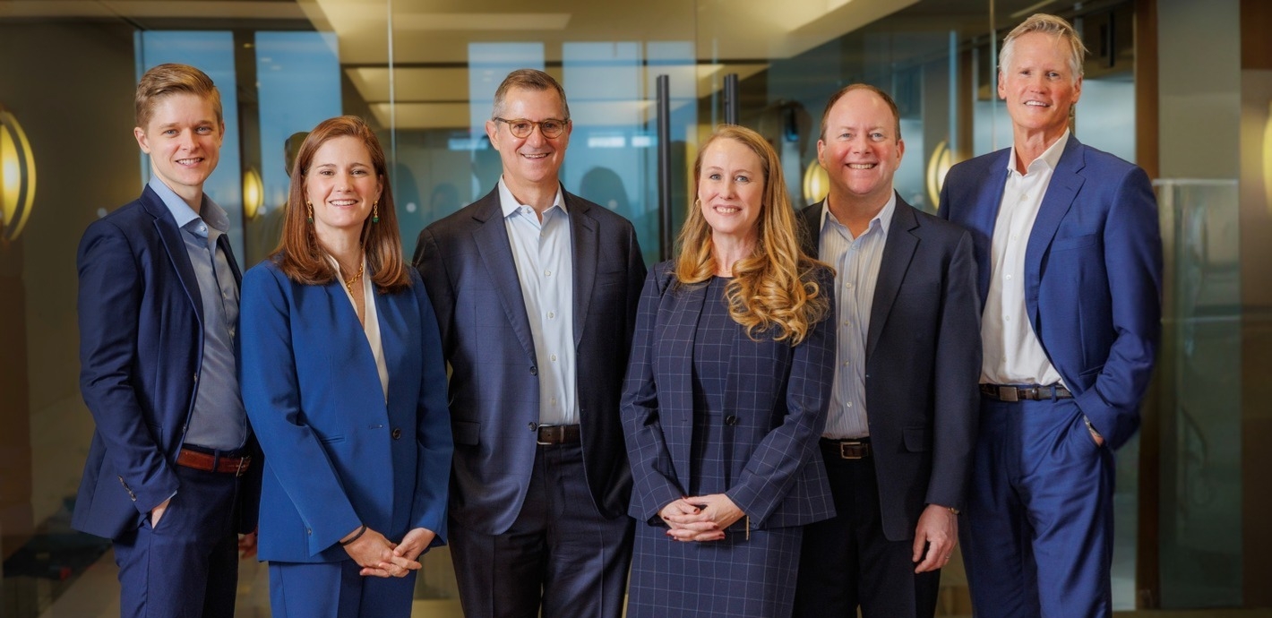 Six professionally dressed people, four men and two women, stand smiling together in an office setting with glass walls and wooden floors. One woman wears boots and a plaid dress; the others wear business suits.