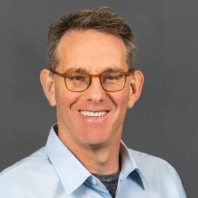 A middle-aged man with short brown hair, wearing glasses and a light blue collared shirt, smiles at the camera against a plain gray background—his confident poise reflects experience at the Wharton School and Theta Equity.