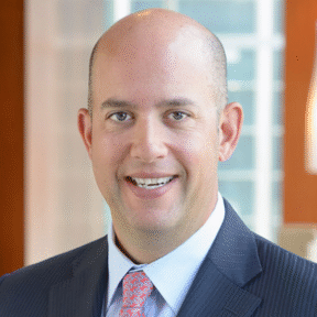 A smiling bald man in a dark pinstripe suit, light blue shirt, and patterned tie stands indoors with blurred Mesirow Financial office windows in the background.