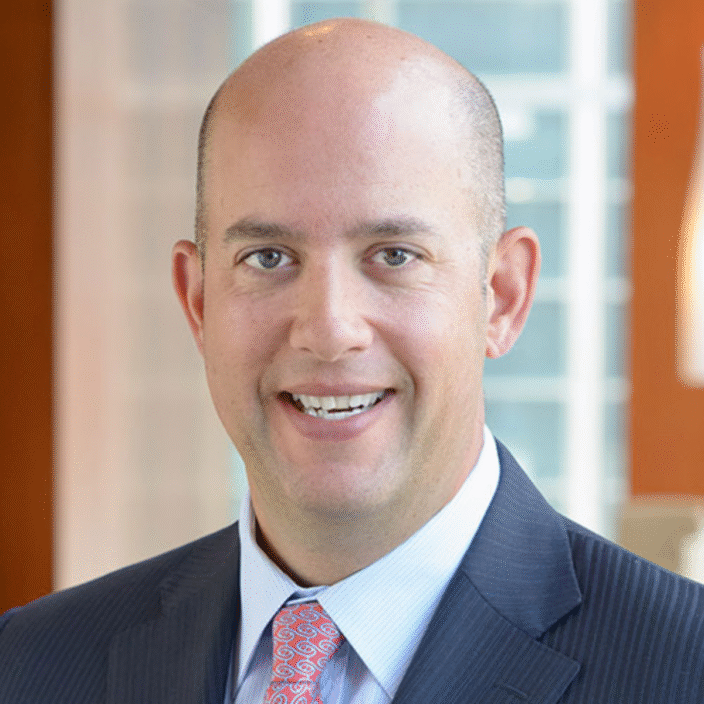 A smiling bald man in a dark pinstripe suit, light blue shirt, and patterned tie stands indoors with blurred Mesirow Financial office windows in the background.
