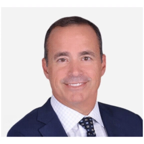A man in a dark blue suit, white checkered shirt, and patterned tie smiles at the camera against a plain white background, representing Capstone Partners with professionalism and confidence.