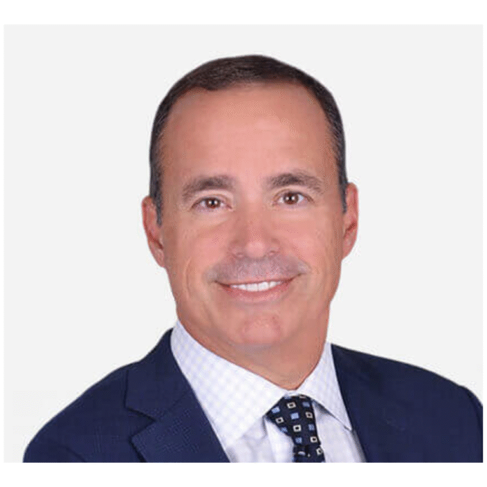 A man in a dark blue suit, white checkered shirt, and patterned tie smiles at the camera against a plain white background, representing Capstone Partners with professionalism and confidence.